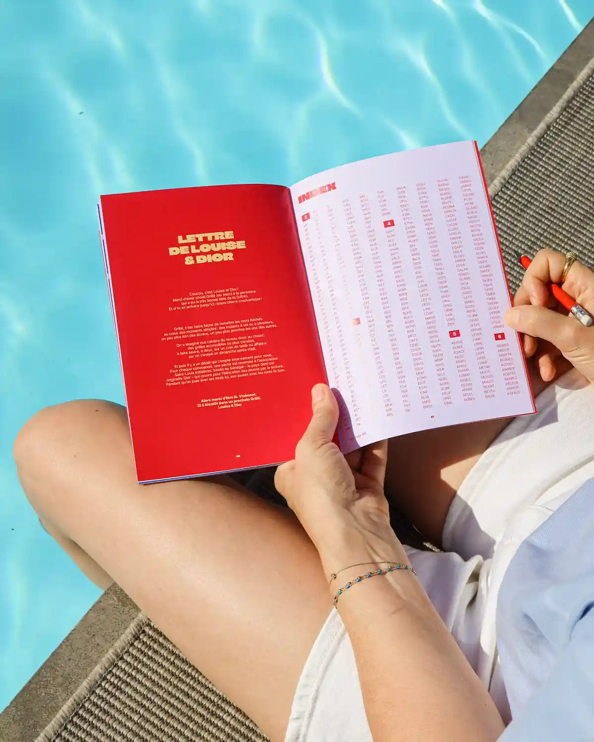 Cahier Grillé TOUT DOUX ouvert au bord d'une piscine, montrant grilles mots fléchés et index coloré en rouge