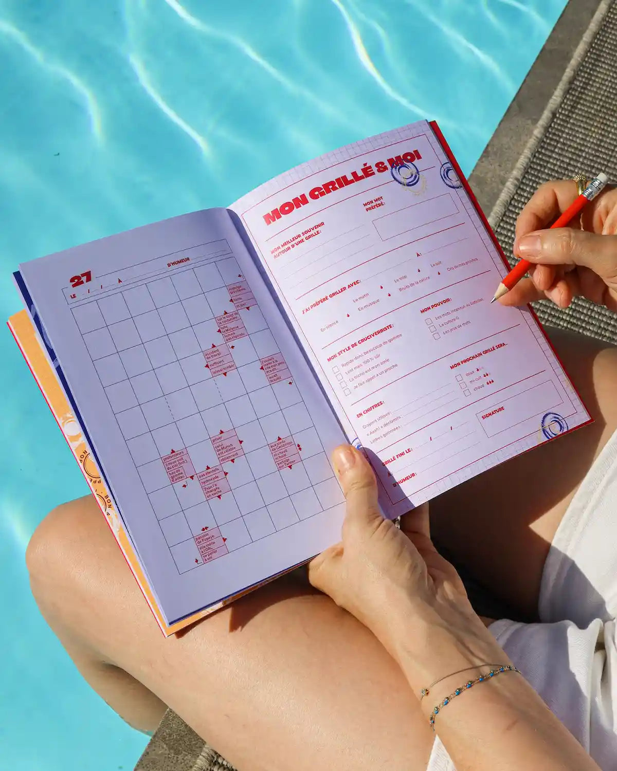Cahier Grillé TOUT DOUX ouvert au bord d'une piscine, montrant grilles mots fléchés colorées et main écrivant avec stylo rouge