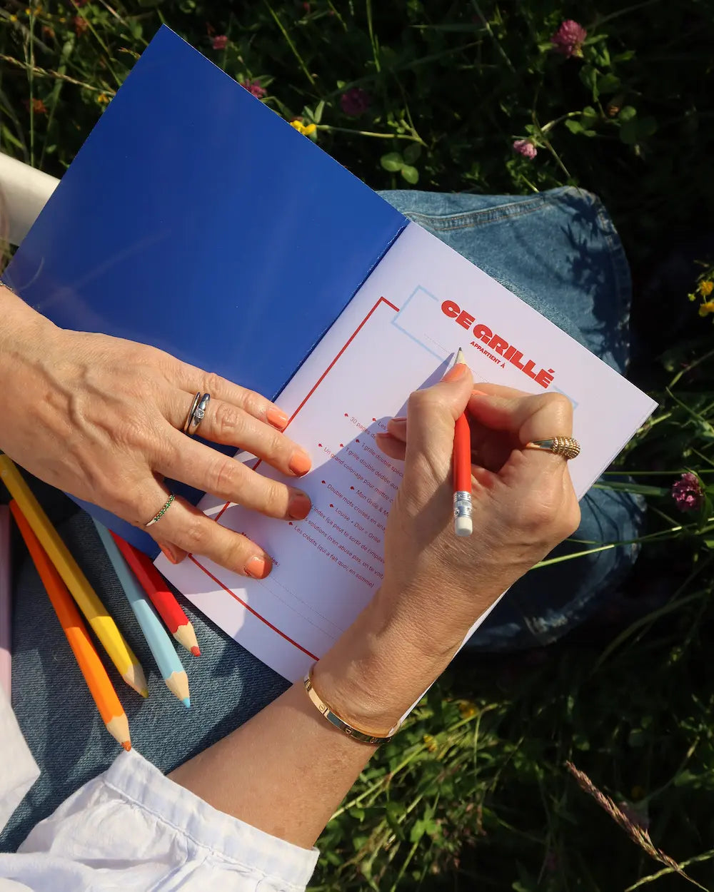Cahier Grillé CULTURE ouvert en couverture bleue, tenu en main avec crayons de couleur, dans un jardin fleuri