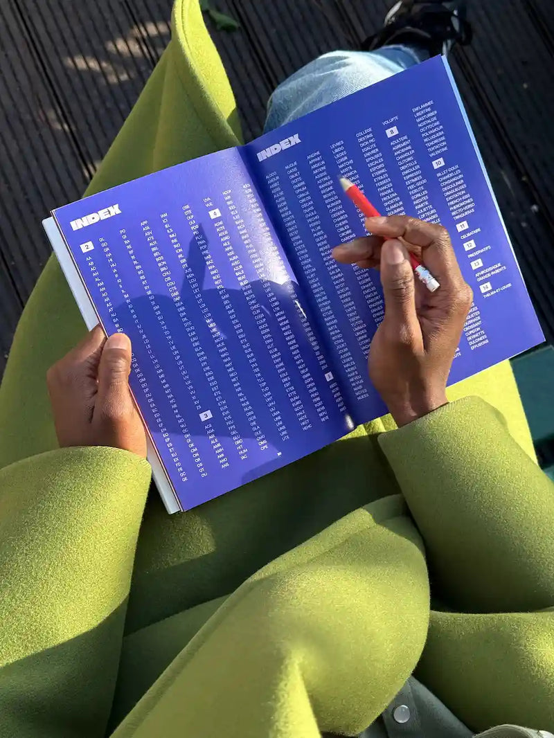 Cahier de mots fléchés Grillé ouvert aux pages index, tenu en main avec un stylo rouge sur fond de grille bleue