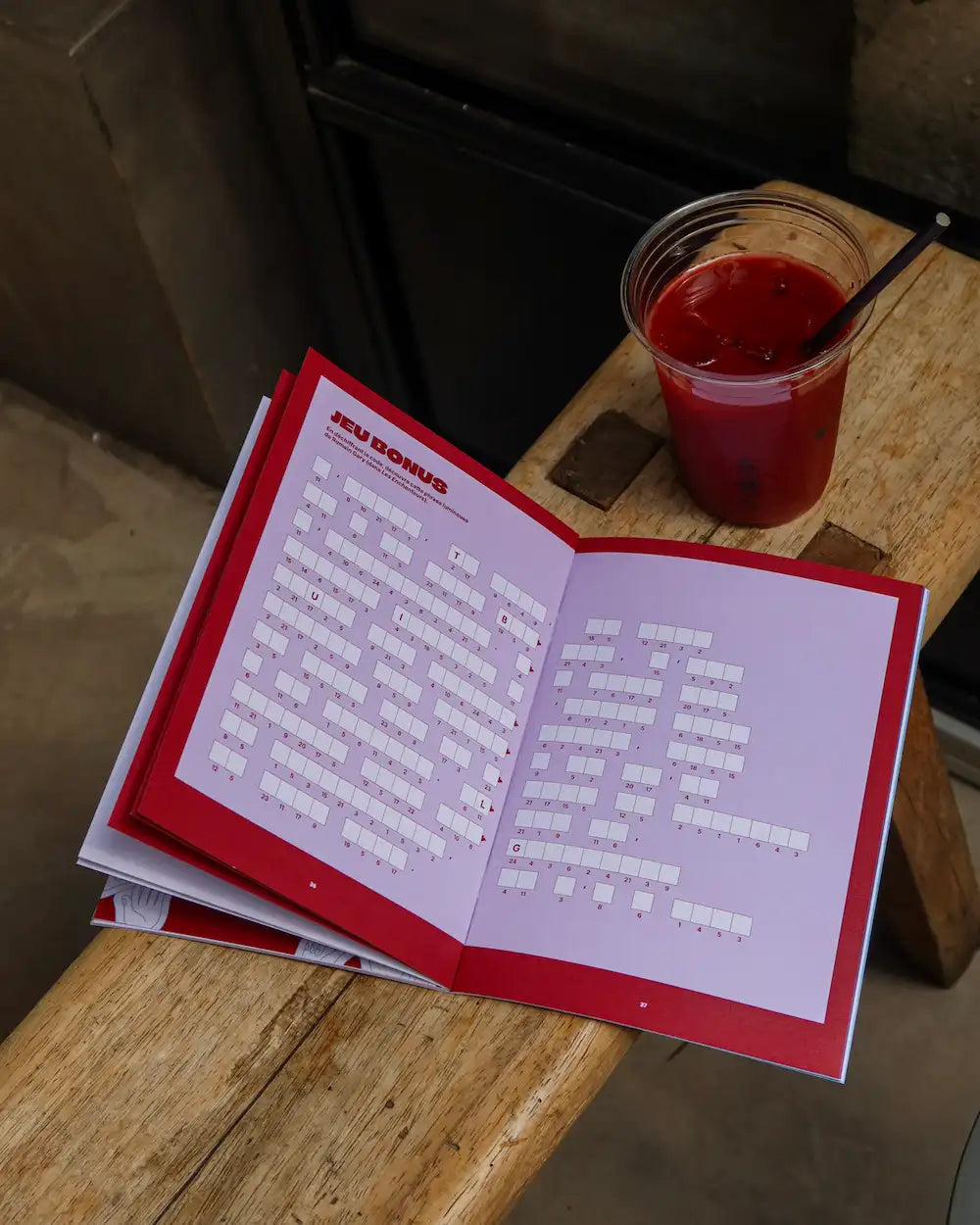 Cahier mots fléchés Grillé Amour avec couverture rouge et intérieur rose, ouvert sur table en bois avec verre de jus
