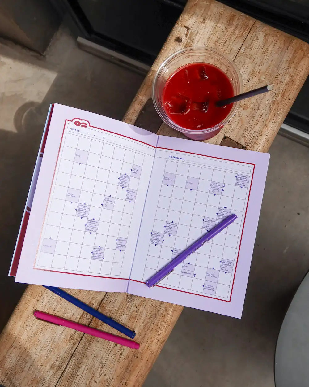 Cahier mots fléchés Grillé Amour ouvert avec stylos colorés et boisson rouge sur table bois