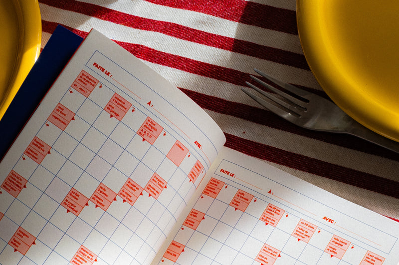 Cahier de mots fléchés Grillé ouvert sur une table dressée avec assiettes jaunes et fourchette, grilles rouge et blanc