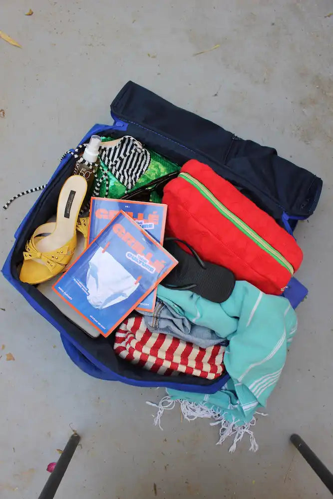 Valise bleue ouverte remplie de vêtements colorés, chaussures jaunes et cahiers de mots fléchés Grillé
