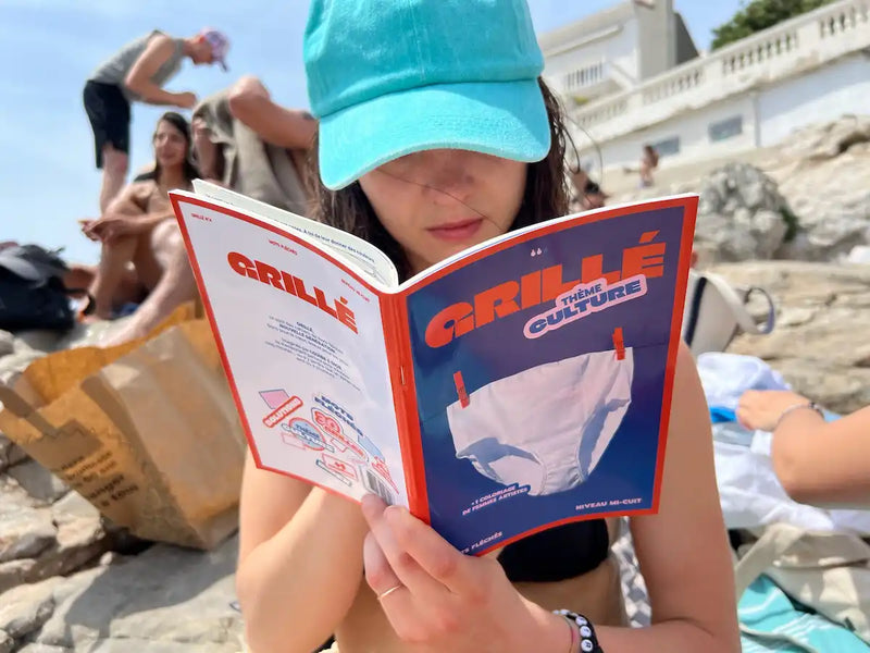 Jeune fille à la plage lisant un cahier Grillé avec couverture bleue et rouge, thème culture pop