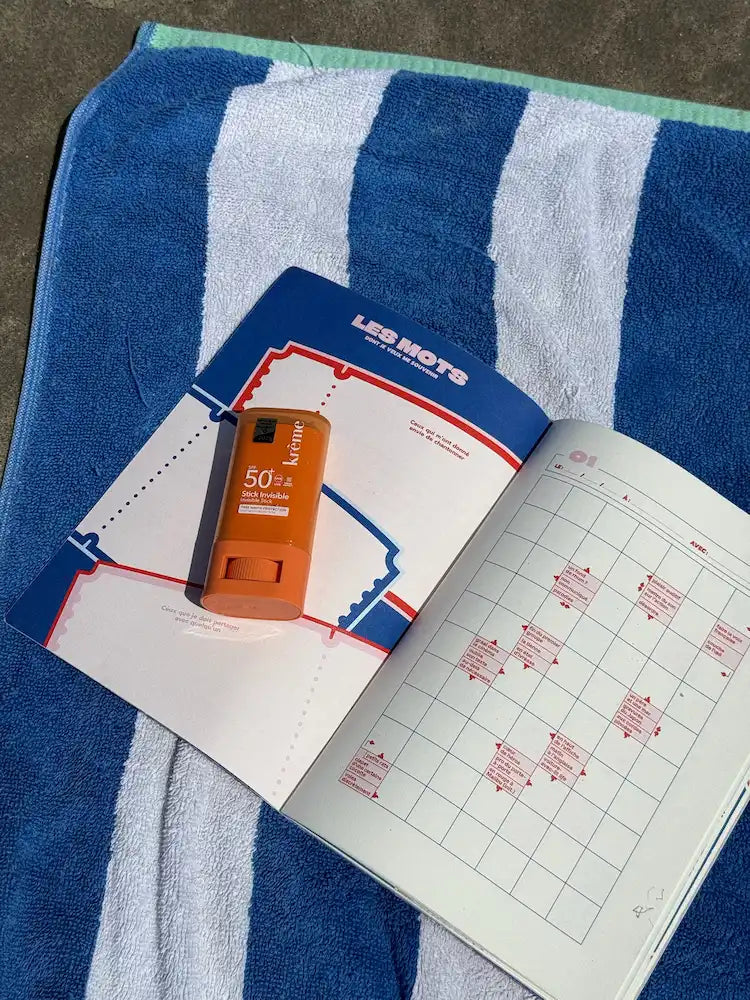 Cahier Grillé mots fléchés rayé bleu blanc avec crème solaire 50+ et calendrier mensuel sur serviette plage